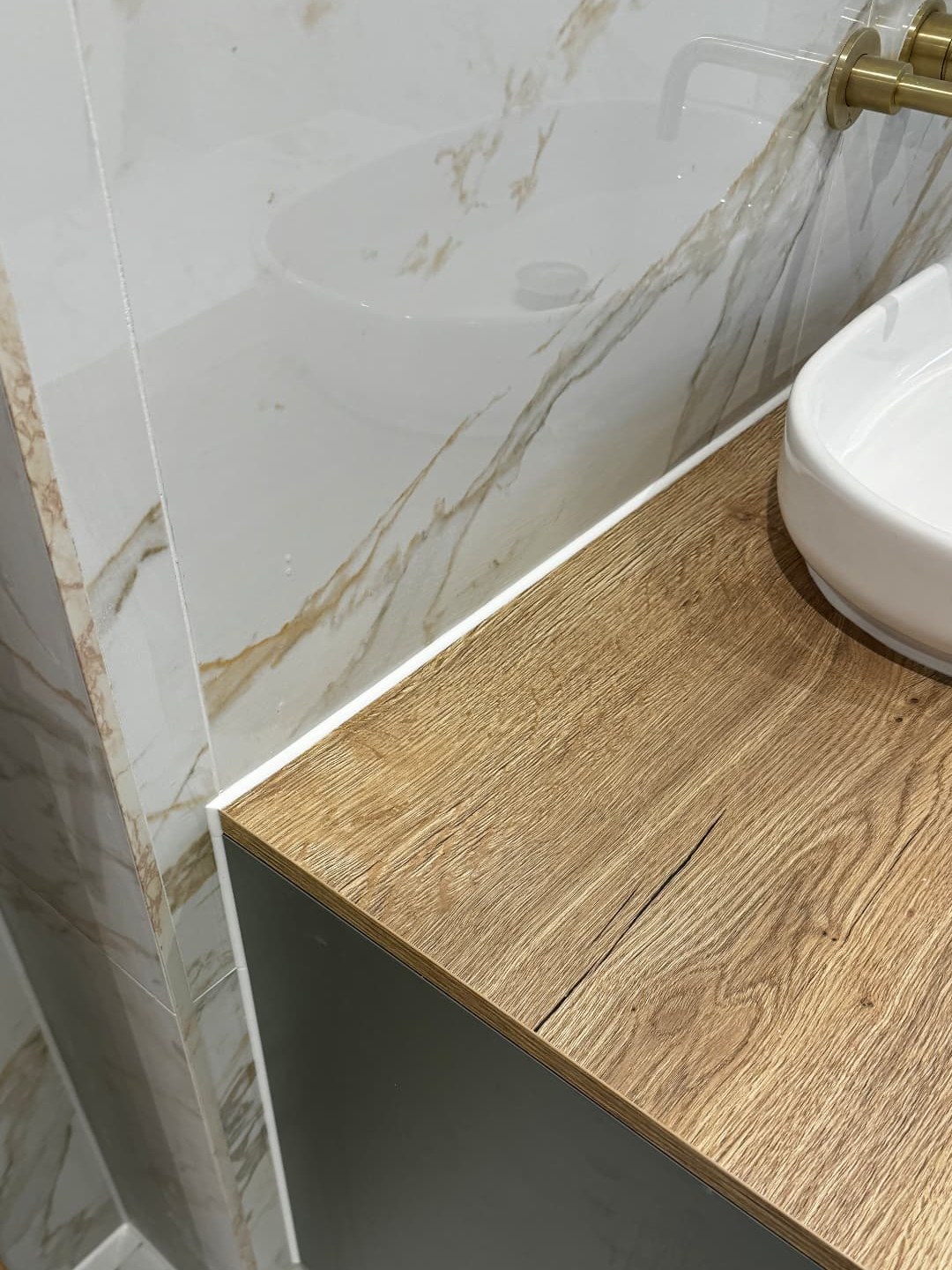 Crisp white silicone sealant lines where marble-effect bathroom tiling meets an oak vanity top with a brushed-gold wall-mounted tap