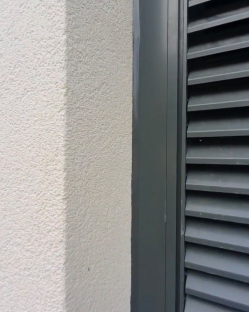 Anthracite mastic joint between rendered wall and a dark aluminium louvred window frame